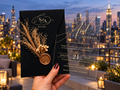 Black monogram envelope with a dried botanical sprig and round wax seal beside a clear gold-lettered invitation, against a rooftop skyline.