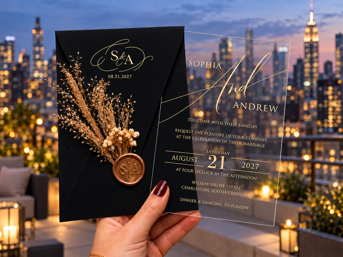 Black monogram envelope with a dried botanical sprig and round wax seal beside a clear gold-lettered invitation, against a rooftop skyline.