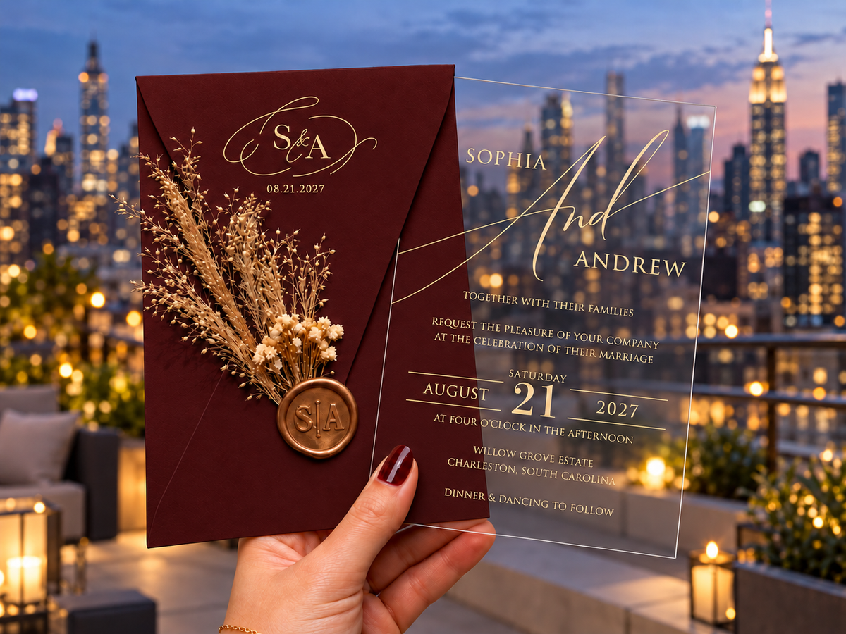 Burgundy monogram envelope with a dried botanical sprig and round wax seal beside a clear gold-lettered invitation, against a rooftop skyline.