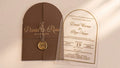 Front view of a brown arched gatefold cover with a printed band, wax seal and dried flowers beside a clear acrylic wedding invitation.