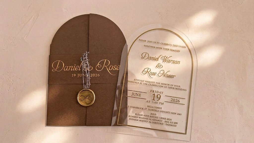 Wide front view of the brown arched cover and clear invitation card side by side, with soft patches of sunlight across the background.