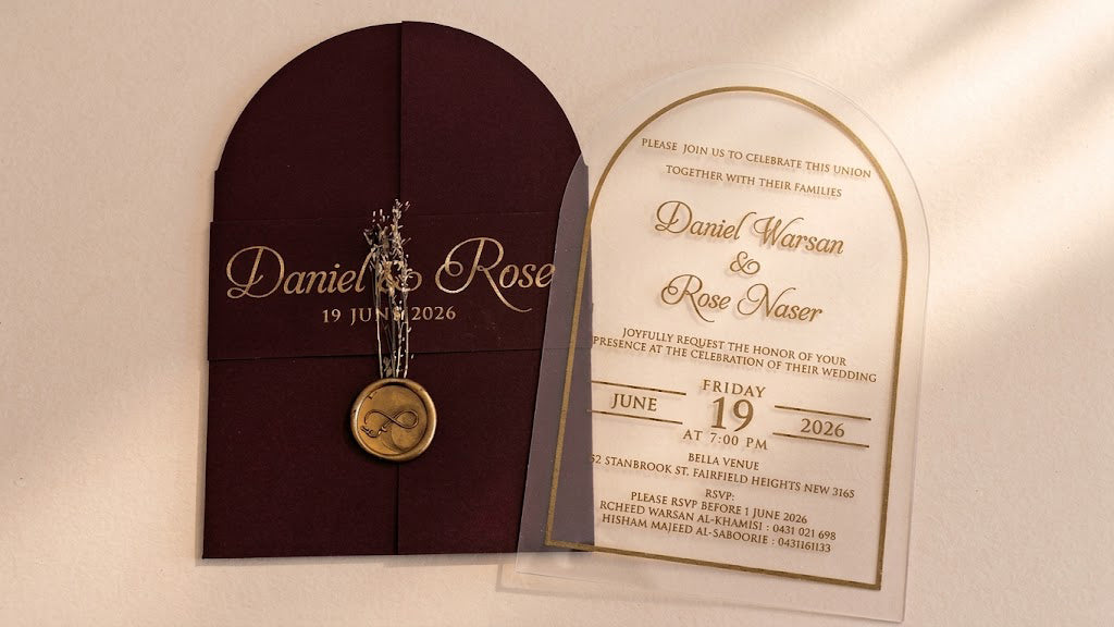 Front view of a burgundy arched gatefold cover with a printed band, wax seal and dried flowers beside a clear acrylic wedding invitation.