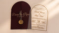 Front view of a burgundy arched gatefold cover with a printed band, wax seal and dried flowers beside a clear acrylic wedding invitation.