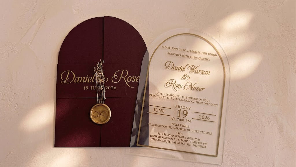 Wide front view of the burgundy arched cover and clear invitation card side by side, with soft patches of sunlight across the background.