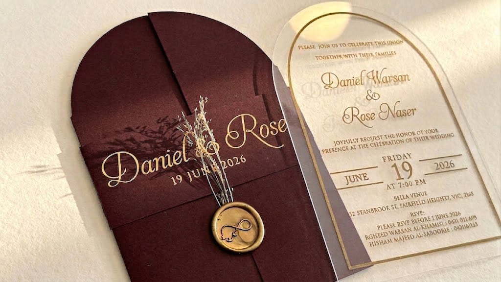 Close view of the burgundy cover's printed paper band, dried floral stems and round wax seal, beside the arched acrylic invitation.