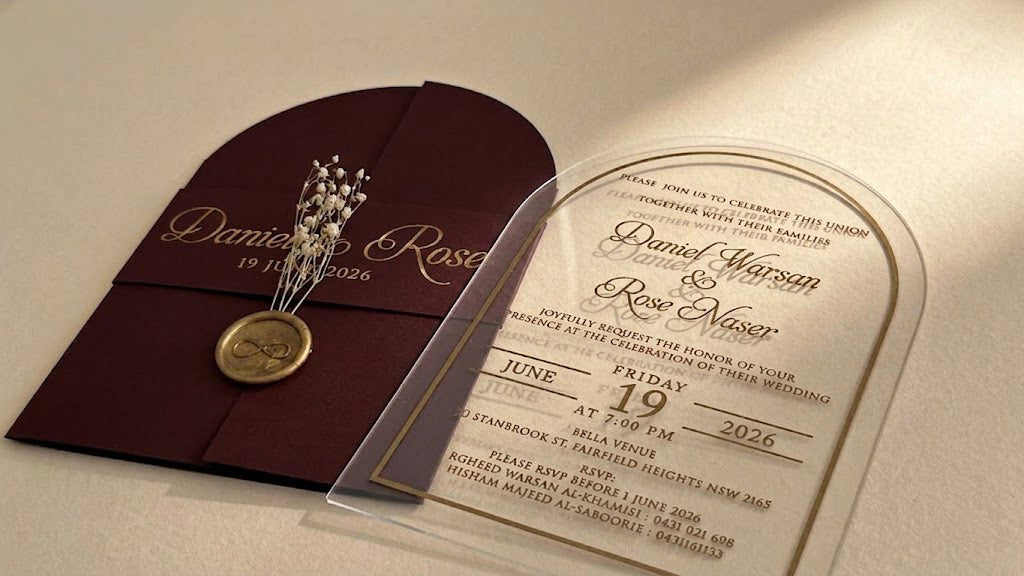 Low view of the burgundy invitation set showing the clear acrylic card's transparent outer edge and lettering beside the closed cover.