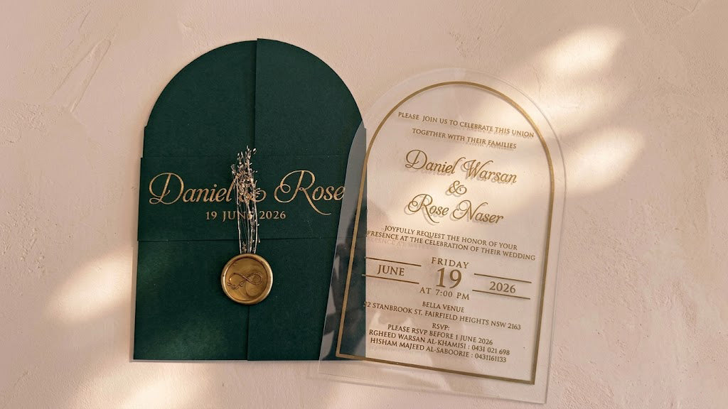 Wide front view of the green arched cover and clear invitation card side by side, with soft patches of sunlight across the background.