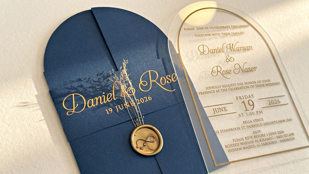 Close view of the navy blue cover's printed paper band, dried floral stems and round wax seal, beside the arched acrylic invitation.