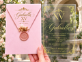 Clear acrylic Quinceañera invitation with a gold ornate frame, pink envelope, pink wax seal and dried flowers.