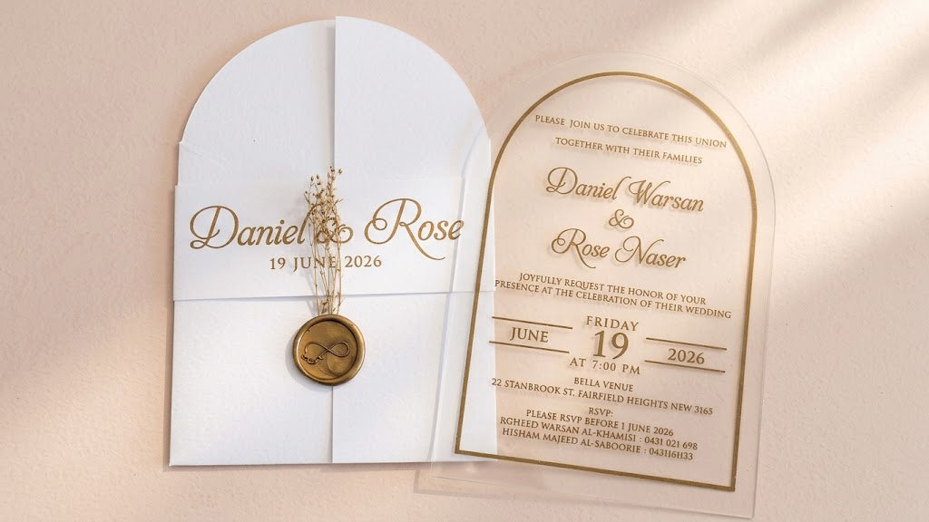 Front view of a white arched gatefold cover with a printed band, wax seal and dried flowers beside a clear acrylic wedding invitation.