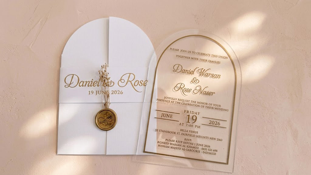 Wide front view of the white arched cover and clear invitation card side by side, with soft patches of sunlight across the background.