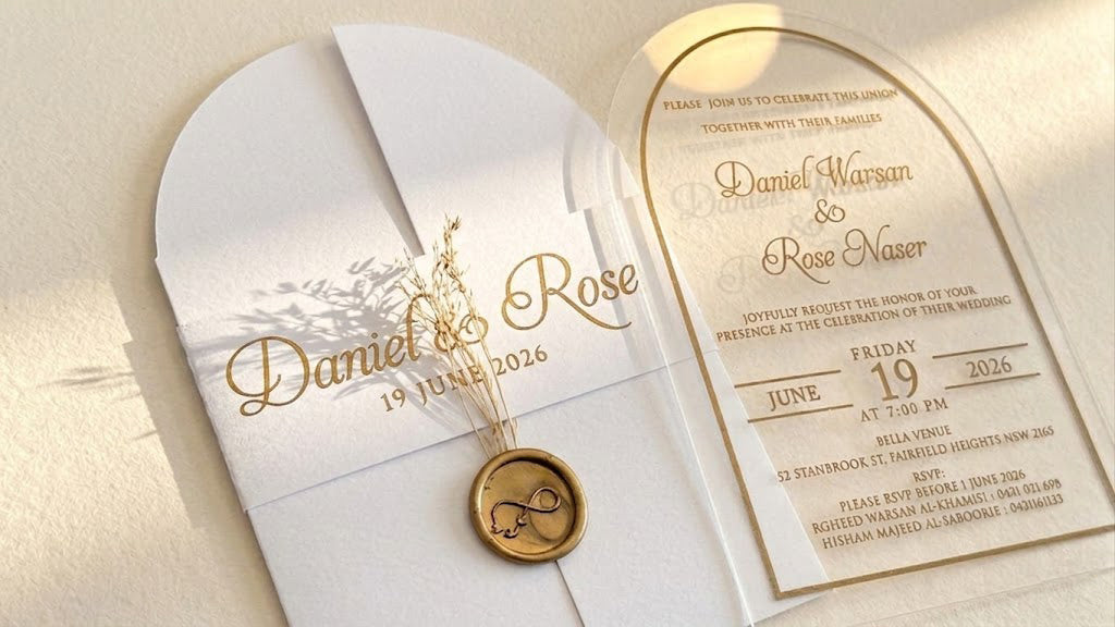Close view of the white cover's printed paper band, dried floral stems and round wax seal, beside the arched acrylic invitation.
