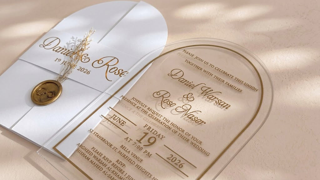 Angled close-up of the clear arched acrylic card's names, date and border, with the white cover, wax seal and dried flowers behind it.
