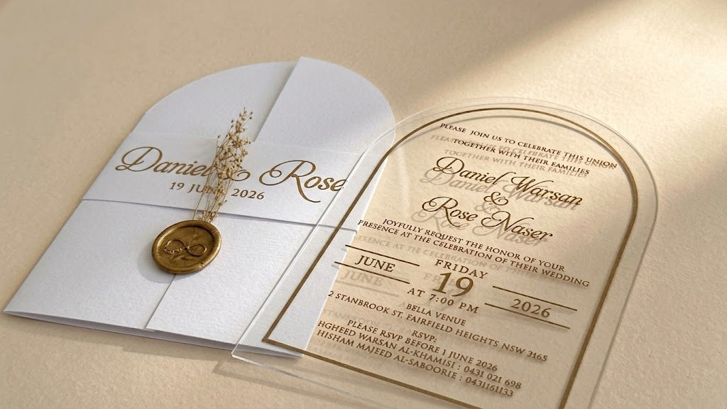 Low view of the white invitation set showing the clear acrylic card's transparent outer edge and lettering beside the closed cover.